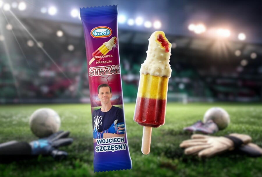 Nowe Lody Koral „STRZAŁ” o smaku truskawki i marakui w polewie, reklamowane przez Wojciecha Szczęsnego na tle stadionu piłkarskiego. Obok opakowania z wizerunkiem i podpisem bramkarza widoczny jest lód na patyku oraz leżące na murawie rękawice bramkarskie i piłki.