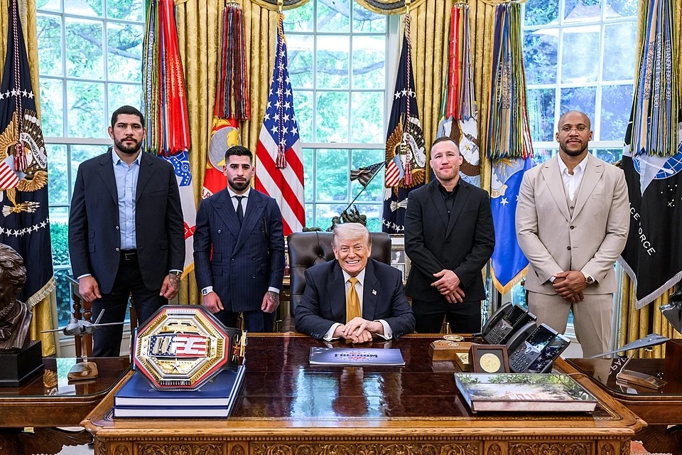Donald Trump siedzący przy biurku w Gabinecie Owalnym z zawodnikami UFC stojącymi obok podczas spotkania w Białym Domu.