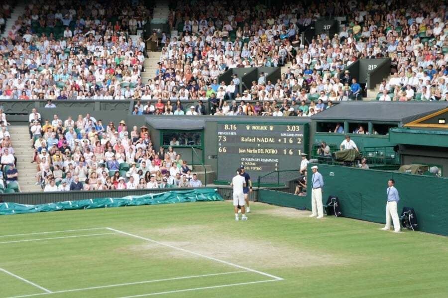 Widok na kort trawiasty podczas prestiżowego turnieju Wimbledon, na którym zawodnicy Rafael Nadal i Juan Martín del Potro stoją przy siatce. W tle widać tablicę wyników Rolex oraz trybuny pełne kibiców.
