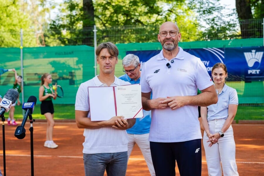 Minister sportu Jakub Rutnicki i przedstawiciele klubu tenisowego prezentują oficjalny dokument dofinansowania na modernizację kompleksów tenisowych podczas konferencji prasowej na korcie.