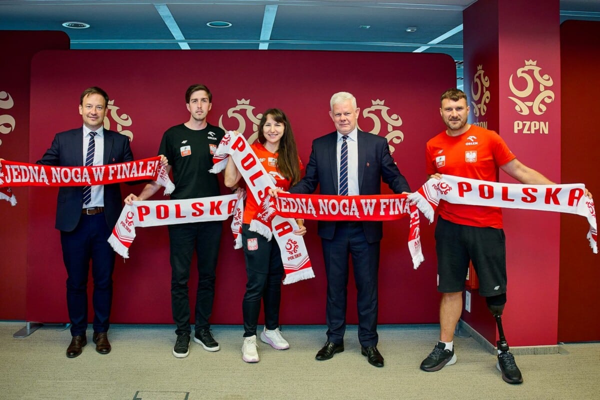 Pięciu przedstawicieli PZPN oraz Polskiego Związku Amp Futbol stoi na tle bordowej ścianki z logo PZPN i trzyma szaliki kibica z napisami Polska oraz Jedna noga w finale.
