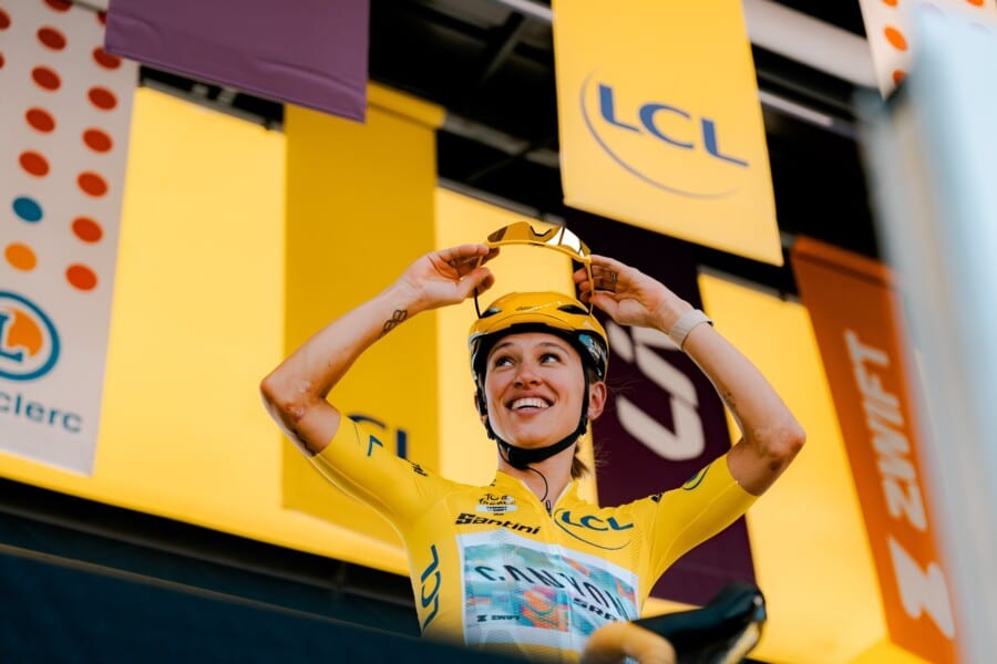 Uśmiechnięta Katarzyna Niewiadoma-Phinney w żółtej koszulce liderki zakładająca okulary kolarskie na podium Tour de France Femmes.