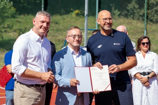 Trzech mężczyzn podczas uroczystości sportowej, jeden z nich trzyma akt powołania, w tle kort i publiczność.
