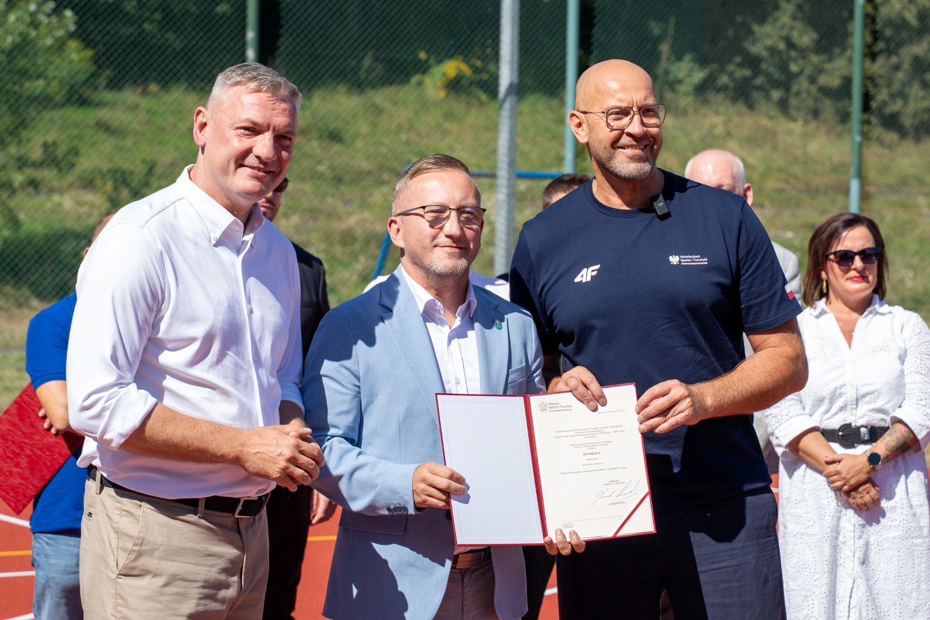 Trzech mężczyzn podczas uroczystości sportowej, jeden z nich trzyma akt powołania, w tle kort i publiczność.