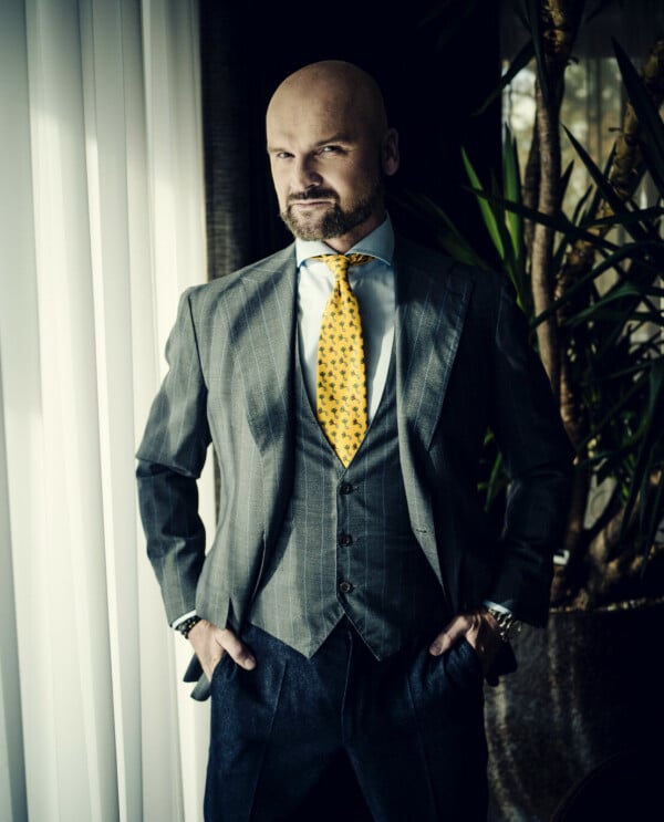 Rafał Brzoska, założyciel i prezes InPostu, ubrany w szary garnitur, kamizelkę i żółty krawat, stojący przy oknie.