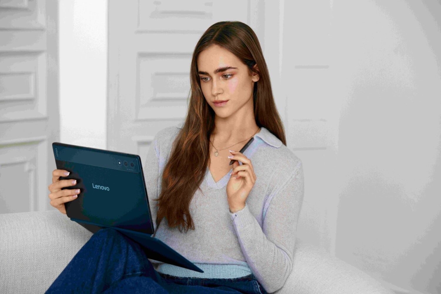 Młoda sprinterka Anastazja Kuś używa tabletu Lenovo Yoga w ciemnoniebieskim kolorze. Siedzi wygodnie i trzyma w dłoni rysik, prezentując urządzenie w jasnym, nowoczesnym wnętrzu. Fotografia promocyjna podkreśla połączenie technologii i stylu życia młodego pokolenia.